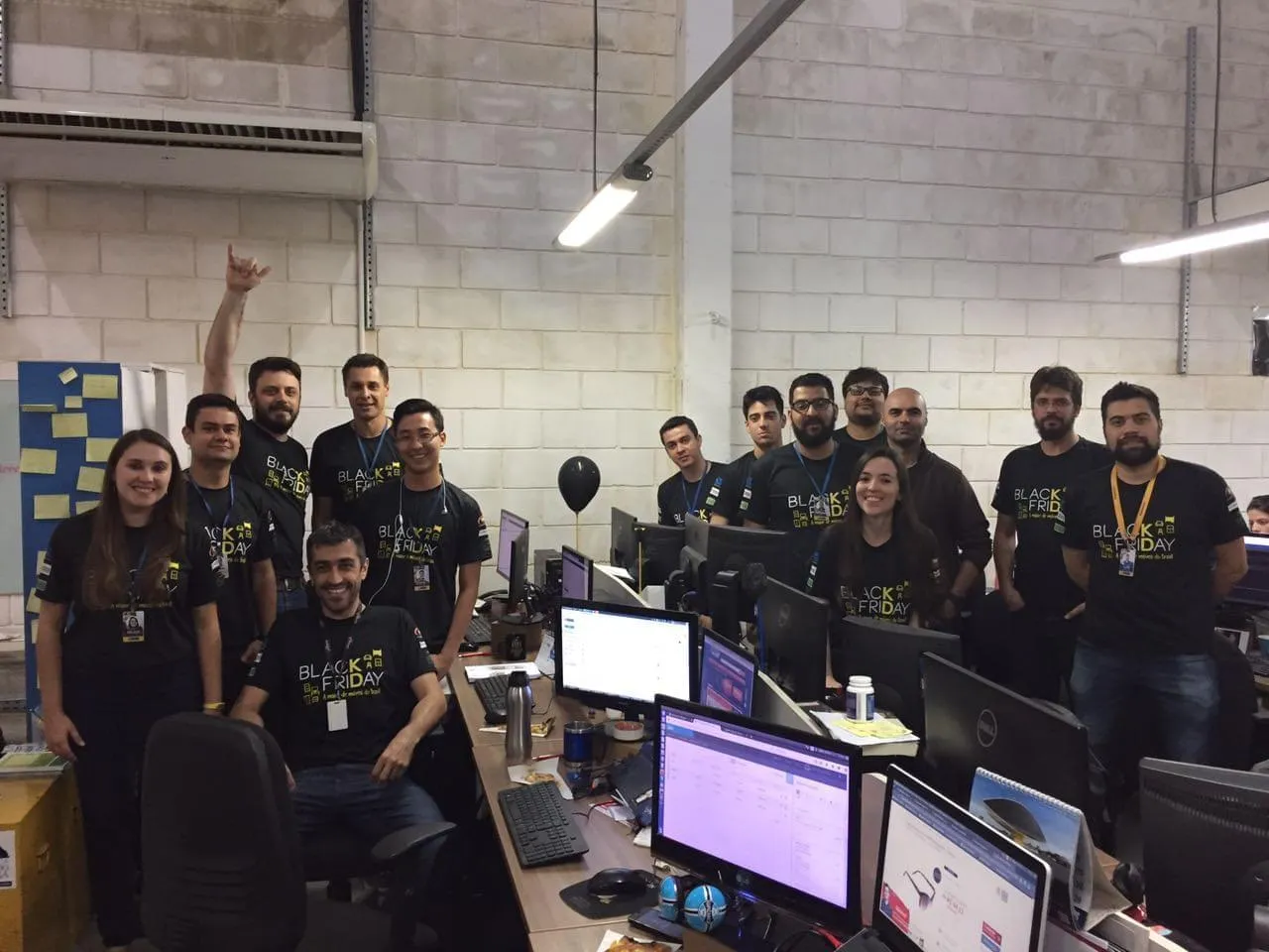 Foto do time de tecnologia posando no escritório, todos de camiseta preta da Black Friday, entre mesas com monitores ligados.