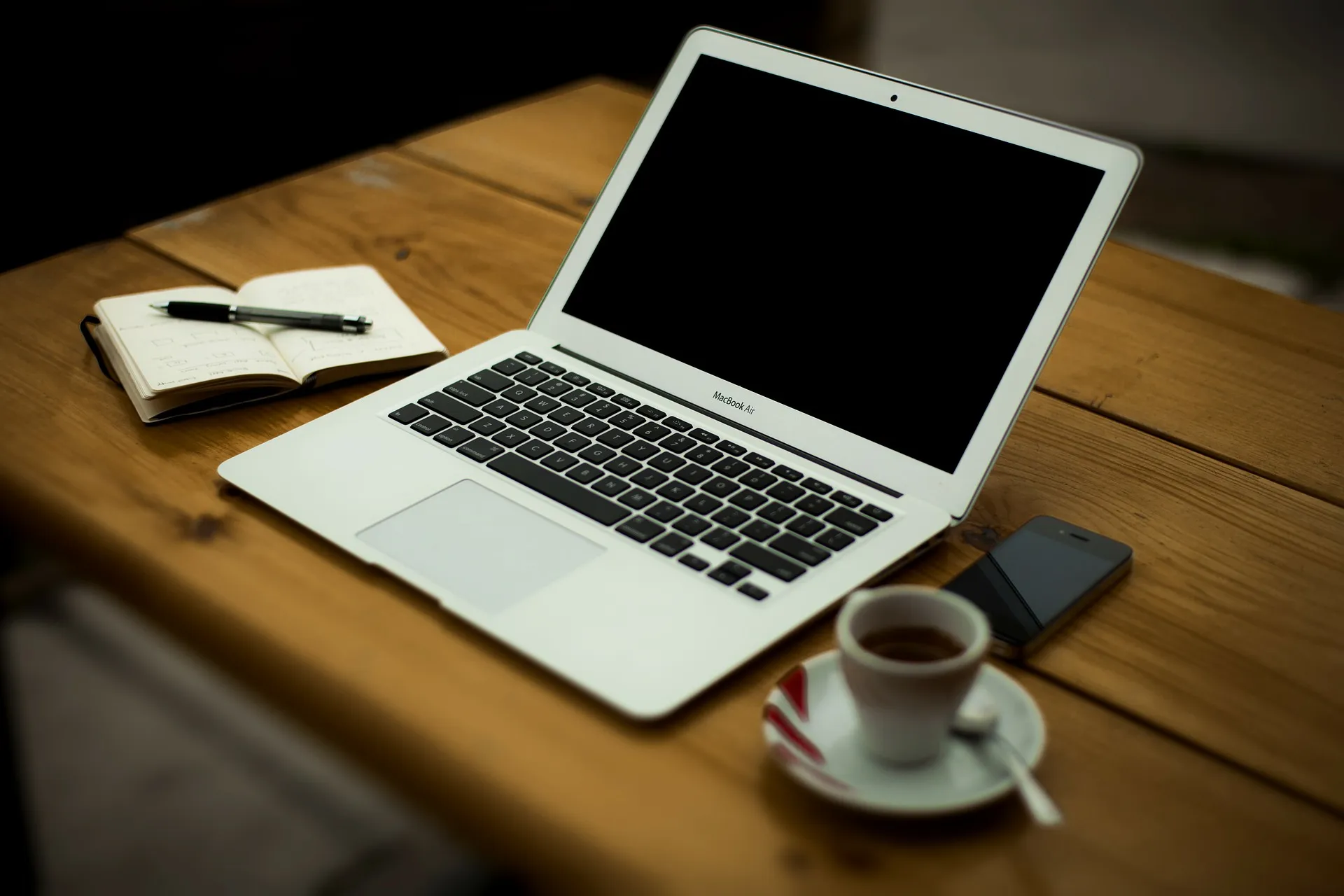 MacBook Air aberto com a tela apagada sobre uma mesa de madeira, ao lado de um caderno com caneta, um celular e uma xícara de café.