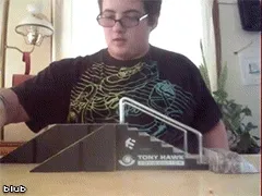 Menino de óculos sentado à mesa, empurrando um skate de dedo por uma rampa de brinquedo.